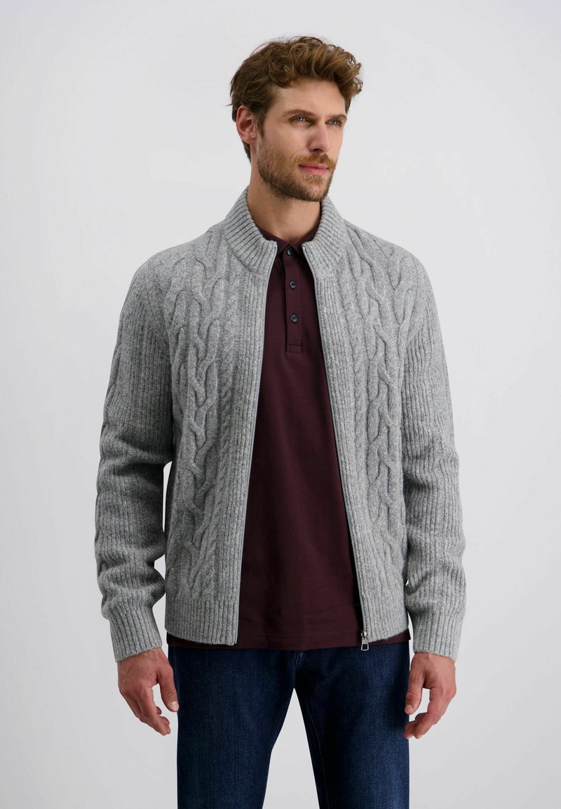 Grau gestrickter Cardigan mit durchgehendem Reißverschluss, gerippten Bündchen und hohem Kragen. Auf der Vorderseite mit einem Zopfmuster versehen, getragen über einem dunkelbraunen Hemd.