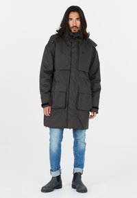 Parka gris foncé avec col montant, fermeture éclair, plusieurs poches avant et coupe décontractée. Portée sur un jean bleu et des bottes noires.