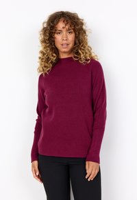 Burgunderfarbener gerippter Strickpullover mit hohem Kragen, langen Ärmeln und lockerer Passform, kombiniert mit schwarzen, enganliegenden Hosen.