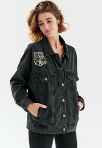 Czarna kurtka dżinsowa z guzikami, dwoma kieszeniami na piersi i haftowanym napisem. Luźny krój, długie rękawy i swobodny design.