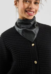 Eine lächelnde Person trägt funkelnde, hängende Ohrringe, einen strukturierten schwarzen Pullover mit goldenen Knöpfen und einen schwarzen Lederschal.
