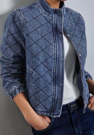 Personne portant une veste matelassée en denim bleu, ouverte sur une chemise blanche, les mains dans les poches d'un jean bleu foncé, sur un fond uni.