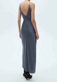 Kobieta ubrana w bezrękawową, bez pleców, sięgającą kostek ciemnoszaro sukienkę z cienkimi ramiączkami i czarnymi szpilkami, stojąca na białym tle.