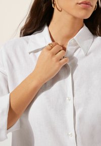 Weiße Leinenbluse mit einem Kragenhals, Knopfleiste und aufgerollten Ärmeln. Zeigt eine Nahaufnahme einer Hand, die einen goldenen Ring trägt.
