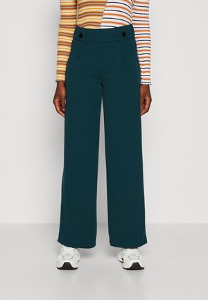 Persoon die donkergroene wijde broek met hoge taille en zwarte knopen draagt, gecombineerd met witte sneakers en een gestreept shirt met lange mouwen.