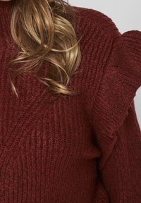 Lockiges, blondes Haar fällt über einen strukturierten, gestreiften, kastanienbraunen Pullover mit einem gerafften Schulterdetail.