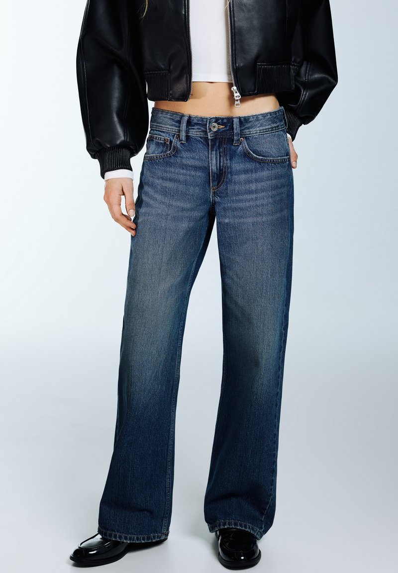 Femme portant un jean bleu à jambes larges, une veste courte noire, un crop top blanc et des mocassins noirs, debout les mains dans les poches.
