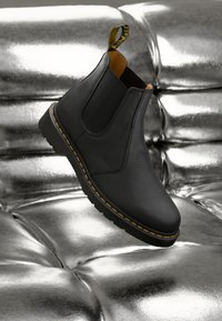 Zwarte leren Chelsea-boot met gele stiksels zwevend boven een glanzend metallic zilveren tufted oppervlak.