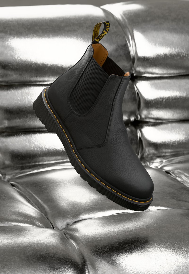 Zwarte leren Chelsea-boot met gele stiksels zwevend boven een glanzend metallic zilveren tufted oppervlak.