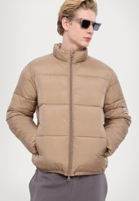 Jachetă puffer de culoare bej deschis, cu guler înalt, design capitonat orizontal, închidere cu fermoar și mansete elastice. Pur tată peste o ținută gri.