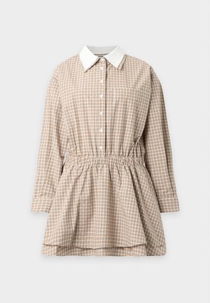 Donna che indossa un mini abito a quadri beige con maniche lunghe, vita raccolta e sandali neri con tacco e cinturini, in piedi davanti a uno sfondo semplice.