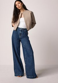 Beige kurz geschnittene Jacke mit Druckknöpfen, weißem Tanktop und hoch taillierten, lässig geschnittenen blauen Jeans. Glatter Stoff und klassische Taschen.