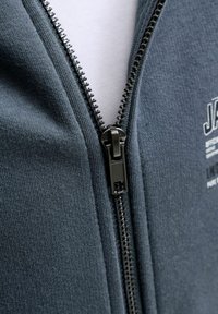 Gros plan d'une fermeture éclair en métal noir partiellement fermée sur un vêtement en tissu gris foncé avec une chemise blanche visible en dessous.