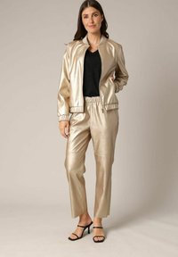 Veste bomber dorée métallique et pantalon assorti avec des poignets et une taille élastiques, associés à un haut noir et des talons noirs à brides.