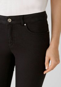Jeans de mezclilla negra con un ajuste slim, que cuentan con un cierre de botón en la parte delantera, dos bolsillos frontales y detalles de costura sutiles. La textura es suave.