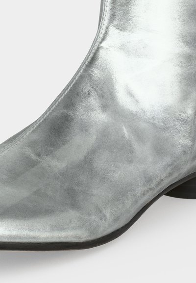 Gros plan d'une botte courte en cuir argent métallisé avec un bout pointu et un petit talon noir sur fond blanc.
