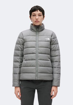 Person iført en lysegrå quiltet North Face jakke med høj krave, stående vendt fremad mod en ensfarvet baggrund.