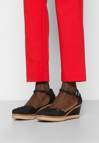 Scarpe con tacco nero in materiale di tela, con cinturino alla caviglia e piattaforma rivestita in juta. Abbinato a pantaloni rossi a tre quarti.