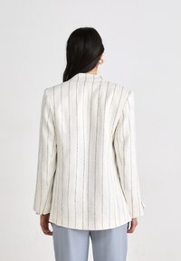Blazer blanco con rayas verticales en navy, corte estructurado, mangas largas y un ligero dobladillo trasero. Usado sobre pantalones azul claro.