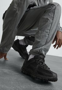 Baskets de sport noires en mesh et matériaux synthétiques, avec une semelle épaisse, un laçage et des accents texturés.