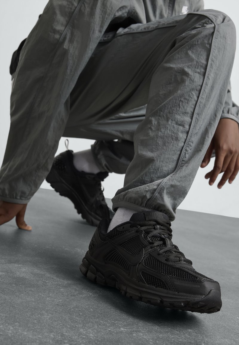 Baskets de sport noires en mesh et matériaux synthétiques, avec une semelle épaisse, un laçage et des accents texturés.