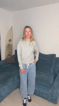 Helehall kapuutsiga pusa, sinised lahtise lõikega teksapüksid ja tumedad tossud. Taustal on sinine diivan ja surfilaud.