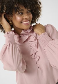 Blusa rosa chiaro con scollo arruffato e dettagli con bottoni. Presenta maniche lunghe a palloncino con polsini arricciati e una texture liscia.