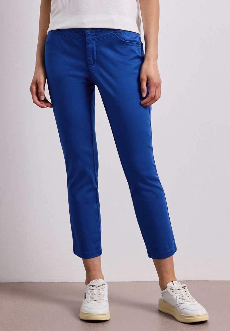 Personne portant un pantalon bleu slim raccourci et des baskets blanches, debout sur un fond gris clair uni.