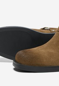 Bottines en daim marron avec semelle en caoutchouc noir, bout rond et détail de boucle sur le côté, sur fond blanc.