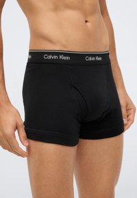Torso masculino vestindo boxers pretos da Calvin Klein com faixa de logo, de pé com os braços relaxados contra um fundo liso.