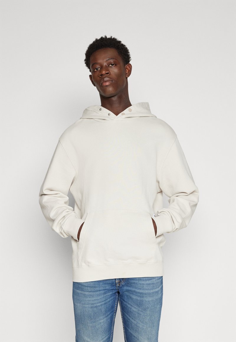 Helle beigefarbene Kapuzensweatjacke mit einer vorderen Tasche, Kordelzug und metallischen Akzenten. Weicher Stoff und ribbelige Bündchen. Getragen mit blauen Jeans.