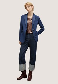 Blazer navy su misura con rever in satin, abbinato a un top di paillettes, jeans in denim scuro con risvolti in denim e stivali con tacco blocco bordeaux.