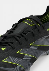 Black football boot featuring textured synthetic material, sleek design, green accents, and black laces. Displays dynamic striped pattern.