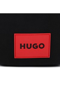 Μαύρο ύφασμα με υφή, που διαθέτει μια έντονη κόκκινη ορθογώνια ετικέτα με ανάγλυφο το λογότυπο «HUGO» σε μαύρα γράμματα.