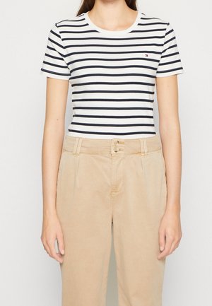 Kobieta w białej koszulce w granatowe paski na krótki rękaw, wpuszczonej w beżowe spodnie z wysokim stanem i podwójnymi guzikami.