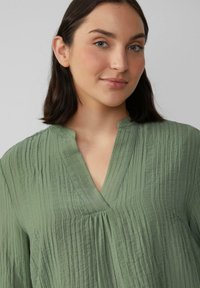 Lichtgroene blouse van een textuurstof met een gekreukt effect. Heeft een V-halslijn en lichte plooien, wat zorgt voor een ontspannen pasvorm.