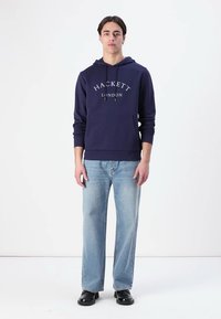 Námořnická mikina s logem "HACKETT LONDON", kapucí na stahování, přední kapsou, spárovaná s světle modrými rovnými džíny a černými botami.