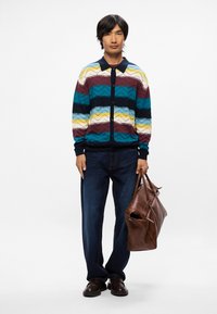 Cardigan en tricot avec des rayures horizontales multicolores, doté d'un col et de boutons navy. Associé à un jean en denim noir et un sac en cuir marron.