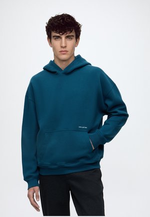 Jeune homme aux cheveux bouclés et foncés, portant un sweat à capuche bleu sarcelle avec poche avant et pantalon noir, debout devant un fond uni.