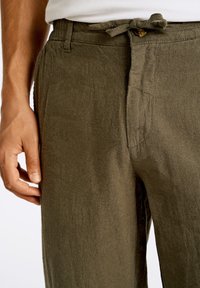 Pantalones de lino ligeros en verde oliva, con cintura ajustable mediante cordón, que presentan una superficie texturizada y bolsillos laterales. Primer plano de la colocación de la mano.