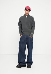 Grijze ritsvest met geribbelde zoom, marineblauwe gestreepte brede broek en een roze pet. De outfit heeft een ontspannen, moderne pasvorm en gestructureerde stof.