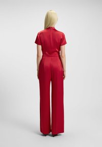 Jumpsuit rosso in satin con maniche corte, colletto alto e chiusura con zip sul retro; presenta gambe larghe e una texture liscia.