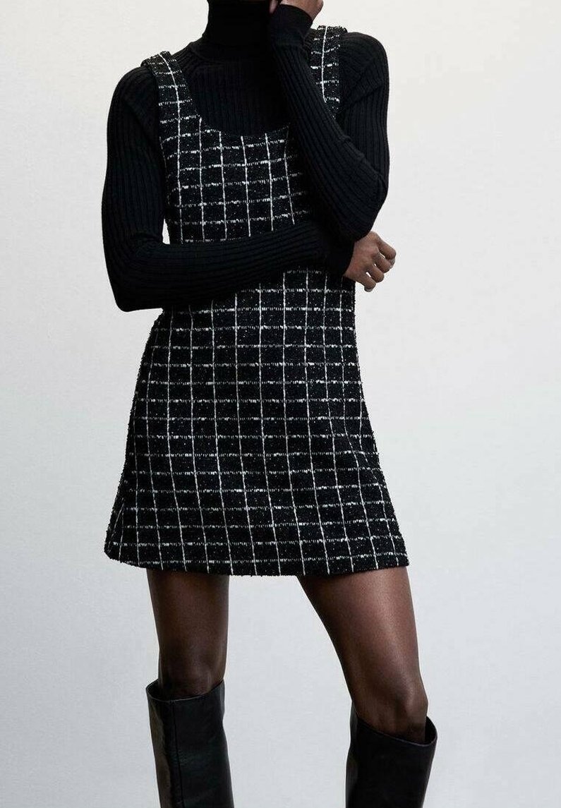 Personne portant un pull à col roulé côtelé noir, une robe sans manches à carreaux noir et blanc, et des bottes noires montantes jusqu'aux genoux sur un fond uni.