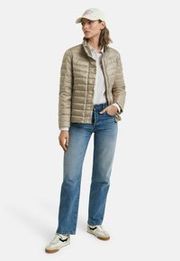 Doudoune beige avec col haut, boutons et texture matelassée, associée à un jean bleu clair et des baskets blanches avec des accents noirs.