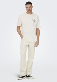 Cream-colored cotton t-shirt features a green graphic on the chest. Paired with light-colored, relaxed-fit pants and black sneakers.