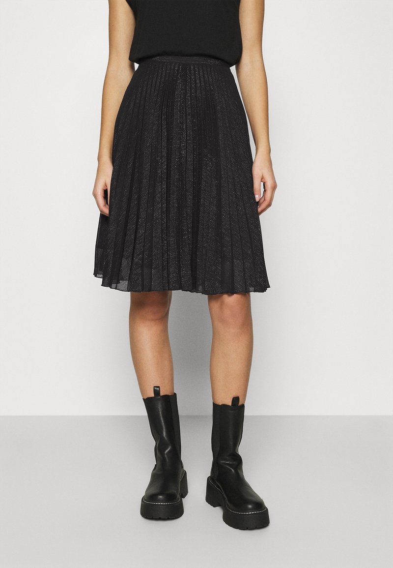 Femme portant une jupe plissée noire longueur genou, un haut noir sans manches et des bottines noires épaisses, debout devant un fond uni.