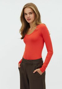 Langsleeve, scoop-neck top in feloranje stof, aangesloten ontwerp. Paar met hoog-gewelfde, donkergroene broeken, handen in de zakken.