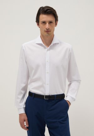 Hombre con cabello castaño vestido con una camisa blanca abotonada y pantalones azules con cinturón negro, de pie con una mano en el bolsillo.