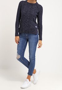 Marinblå stickad tröja med texturerad kabeldesign, kombinerad med ljusblå slitna skinny jeans och vita sneakers.