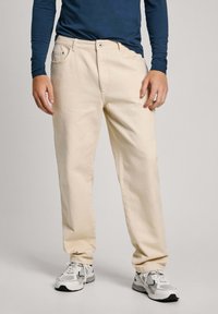 Pantalones de algodón beige con un corte recto, que cuentan con bolsillos frontales y cierre de botón, combinados con zapatillas deportivas en gris y blanco.